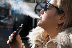 一個戴著太陽眼鏡、身穿淺色外套的人，白天在戶外手持電子香煙時呼出的水蒸氣。. 電子煙 vape