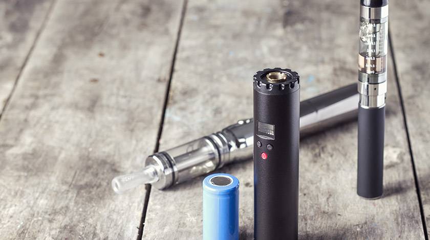 三個 Vape 裝置和一個藍色的圓柱形電池放在一個風化的木頭表面上。它們的大小和設計各不相同，其中一個具有數位顯示和按鈕控制功能。. 電子煙 vape