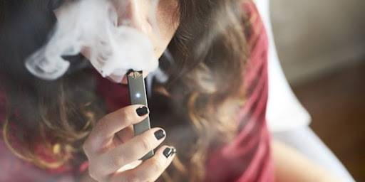 一個留著長髮、指甲塗成黑色的人，正拿著一支 Vape 筆靠近嘴邊吐氣。厚厚的水蒸氣遮住了他們的部分臉部。. 電子煙 vape