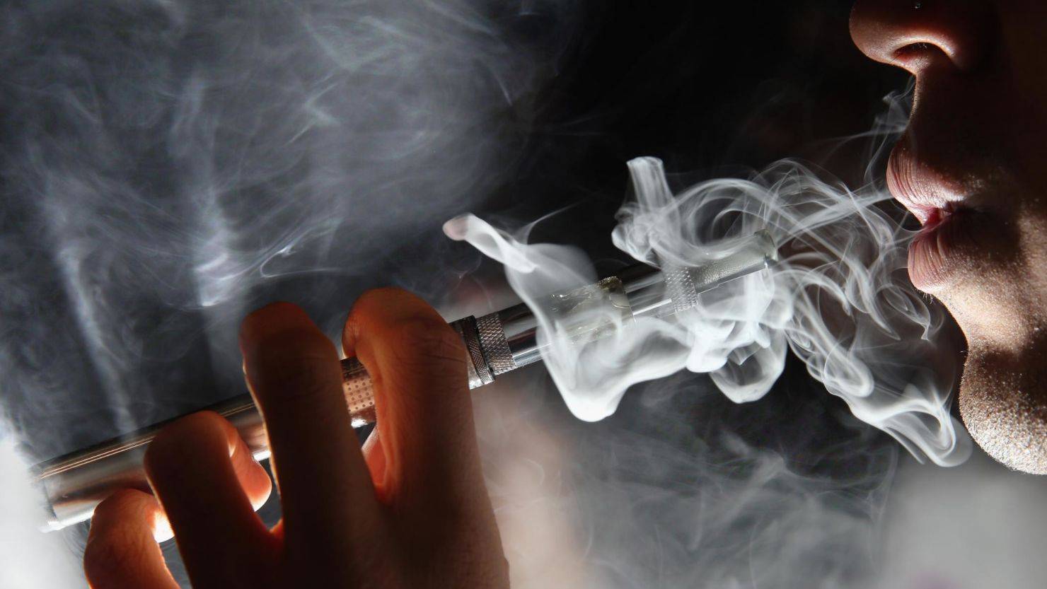 特寫：一個人的嘴從電子香煙中吐出濃稠的蒸氣，在黑暗的背景下有旋轉的煙雲。人物的手拿著吸煙裝置靠近嘴唇。. 電子煙 vape