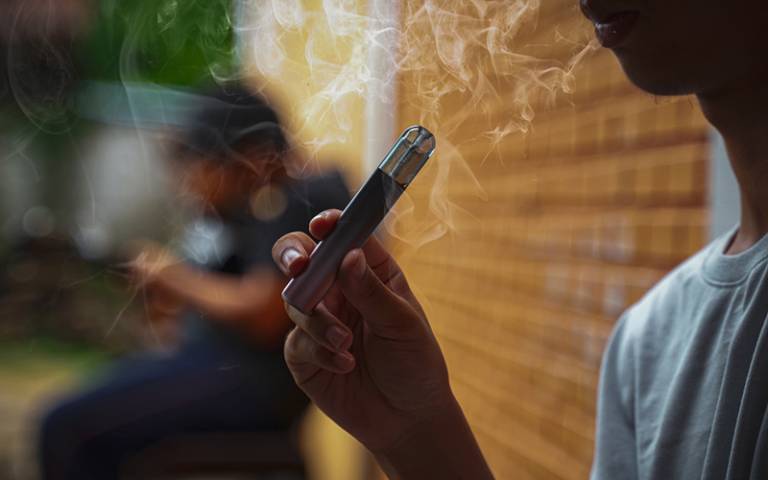 一個人拿著一支煙霧繚繞的 Vape 筆，另一個人坐在磚牆旁的模糊背景中。. 電子煙 vape