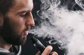 一位鬍鬚男在黑暗背景下使用黑色電子 vape 裝置時呼出一大團蒸汽的特寫。. 電子煙 vape