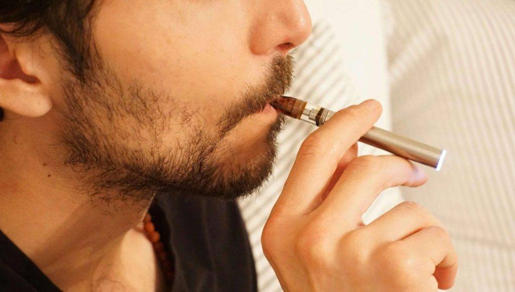 一個留著鬍子的人將電子香煙（vape）放在嘴邊，似乎正在吸入的特寫。背景失焦。. 電子煙 vape
